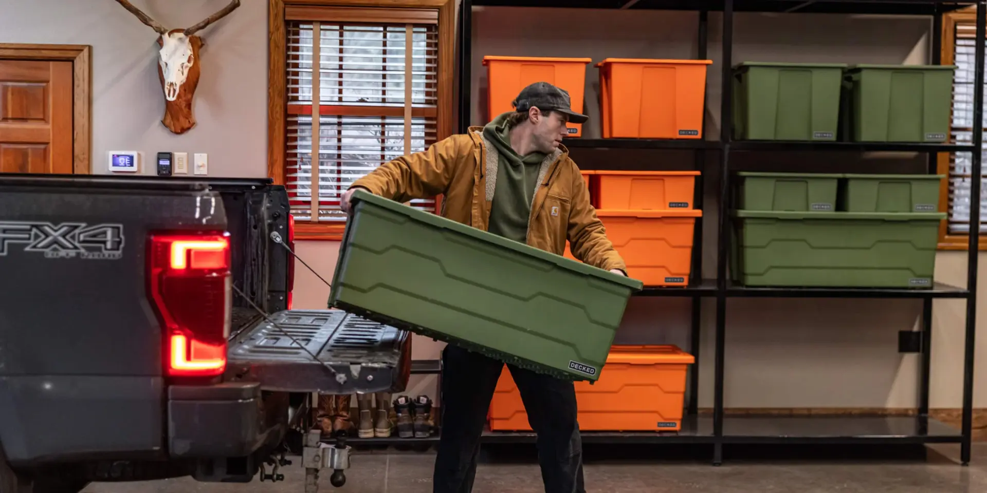 Man loading a green DECKED Payloader bin into a pickup truck in front of shelves of stacked orange and green bins
