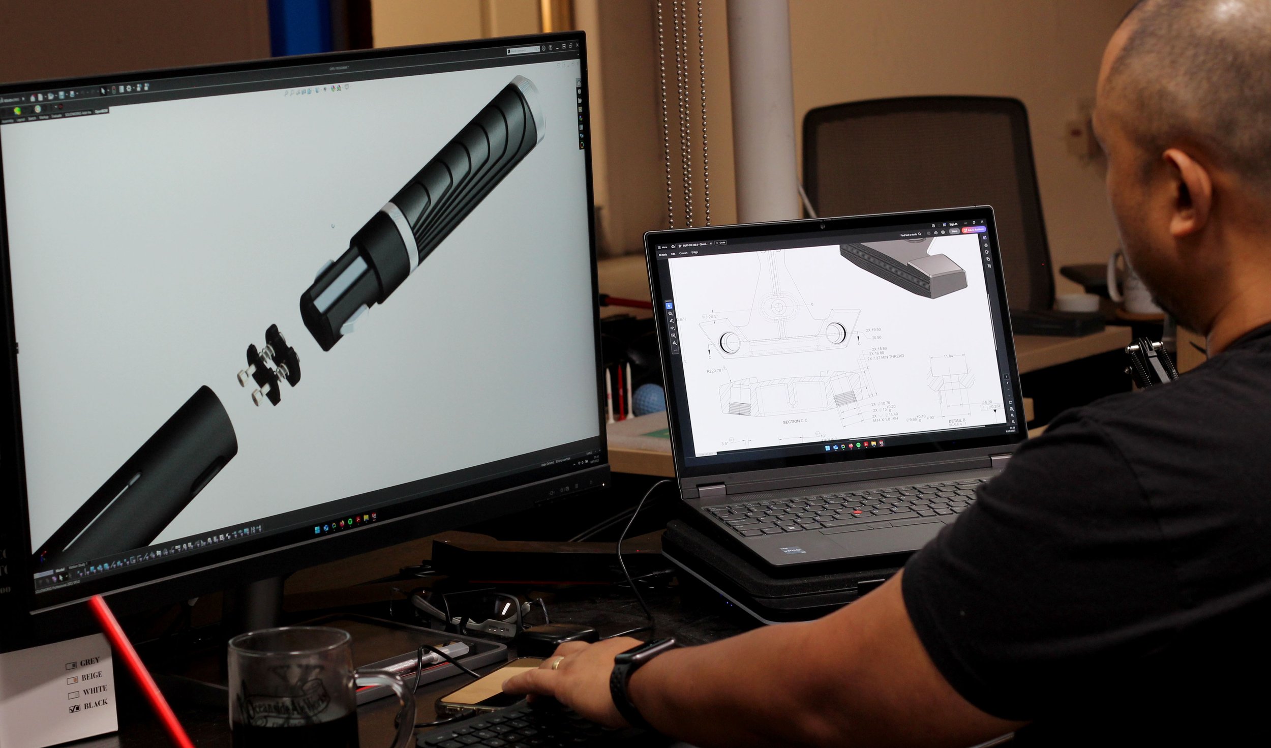 A person at a desk with two monitors showing a 3D model and technical drawings.