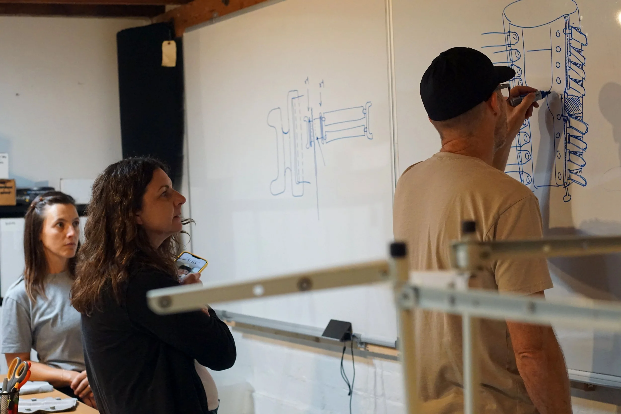 Three people observing a man drawing mechanical diagrams on a whiteboard.